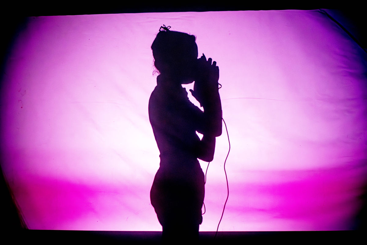 The dancer Victoria Henneberg as a shadow portrayal. She has her hands in front of her face, she stands upright, cables hanging down from her.