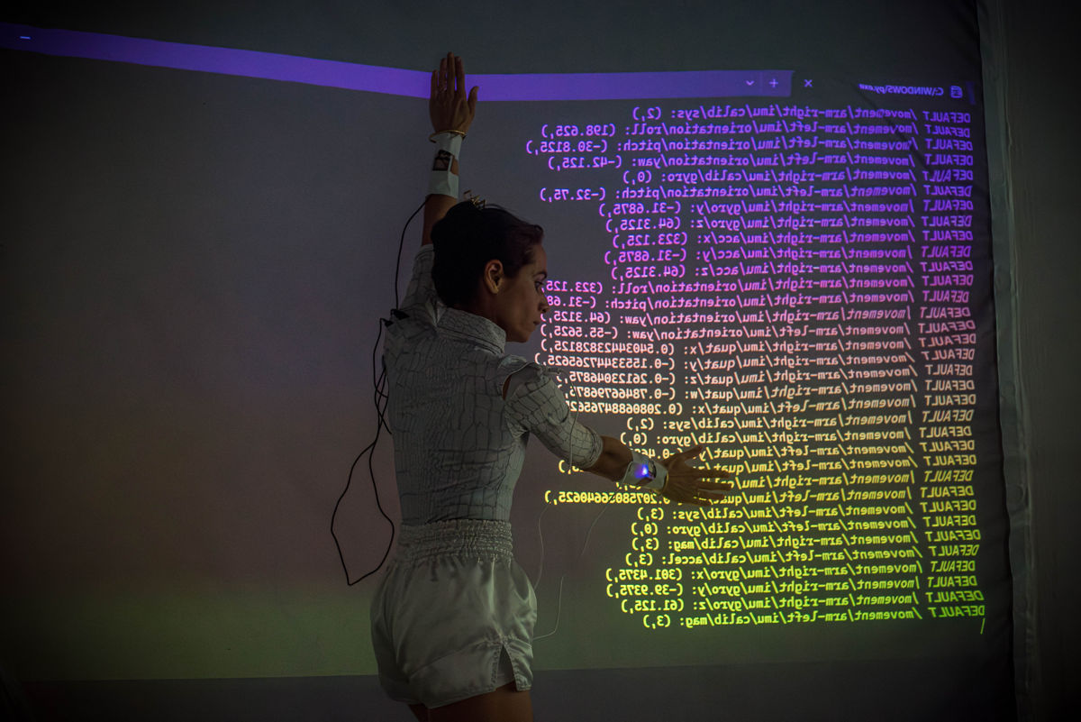 The dancer Victoria Henneberg standing in front of a projection. The projection shows some kind of technical data lines. Her left arm is pointing upwards, her right arm is outstretched at upper body height. She is touching the data with both palms. The cables are still attached to her. They are connected to sensors on the pulse of the arm-hand transition.