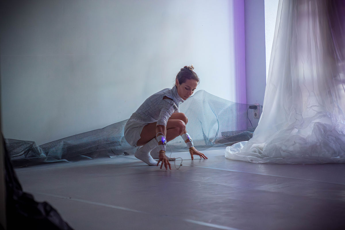 The dancer Victoria Henneberg touches the floor, which is a dance floor, with the fingertips of two hands, sitting in a squatting position.